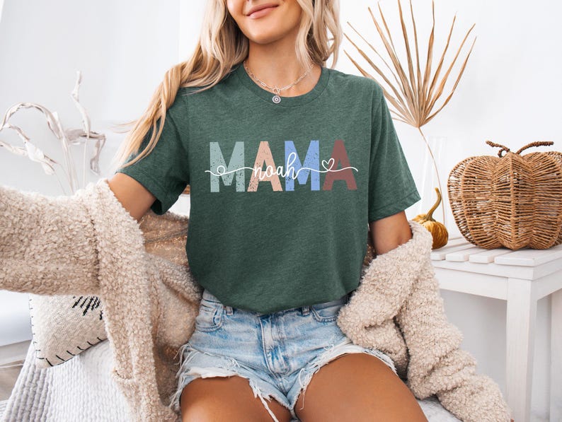 May include: A dark green t-shirt with the word "MAMA" in pastel colors and the name "Noah" in a cursive font. A small heart is next to the name. The shirt is worn with denim shorts and a cream-colored cardigan.