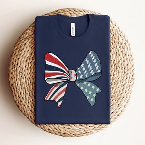 May include: A navy blue t-shirt with a red, white, and blue bow design. The bow is made of fabric with a stars and stripes pattern.