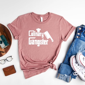 May include: A pink t-shirt with the text "Culinary Gangster" in white, featuring a chef's hat and a butcher knife graphic. The shirt is laid out on a white surface with a wooden cutting board, a camera, glasses, a wallet, blue jeans, and white sneakers.
