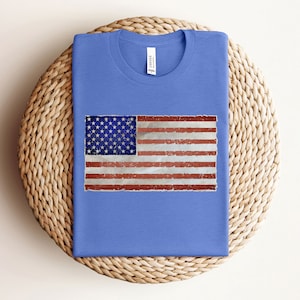 Pode incluir: Uma camiseta azul com um design de bandeira americana desbotada estampado na frente.