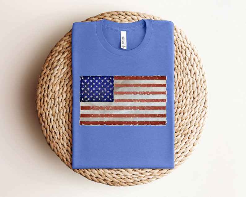 Pode incluir: Uma camiseta azul com um design de bandeira americana desbotada estampado na frente.