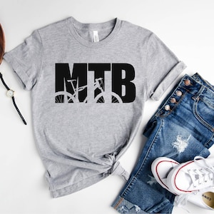 Puede incluir: Una camiseta gris con la palabra "MTB" en letras negras, con una silueta de una bicicleta de monta&ntilde;a dentro de las letras.