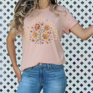 May include: A pink t-shirt with a floral design and the text "Grow Positive Thoughts" in a retro font.