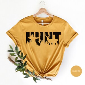 May include: Mustard yellow t-shirt with a black silhouette of a hunter and deer, forming the word "HUNT".