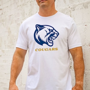 K&ouml;nnte beinhalten: Wei&szlig;es T-Shirt mit einer blauen und wei&szlig;en Grafik eines Pumakopfes und dem Wort "COUGARS" in goldenen Buchstaben.