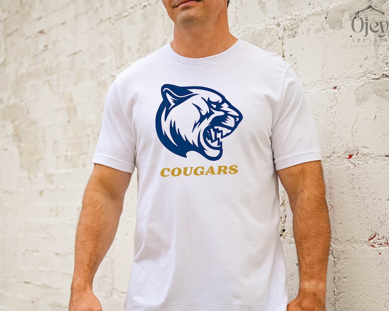K&ouml;nnte beinhalten: Wei&szlig;es T-Shirt mit einer blauen und wei&szlig;en Grafik eines Pumakopfes und dem Wort "COUGARS" in goldenen Buchstaben.