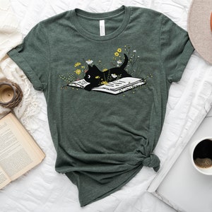 Puede incluir: Una camiseta verde con un gato negro leyendo un libro rodeado de flores amarillas. El texto "HEATHER FOREST" est&aacute; en un c&iacute;rculo blanco en la parte superior izquierda de la imagen.