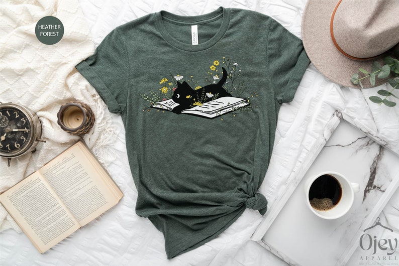 Puede incluir: Una camiseta verde con un gato negro leyendo un libro rodeado de flores amarillas. El texto "HEATHER FOREST" est&aacute; en un c&iacute;rculo blanco en la parte superior izquierda de la imagen.