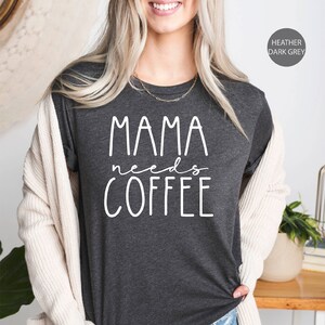 May include: A dark heather grey t-shirt with the text "Mama needs coffee" in white lettering. The shirt is being worn by a person with long blonde hair.