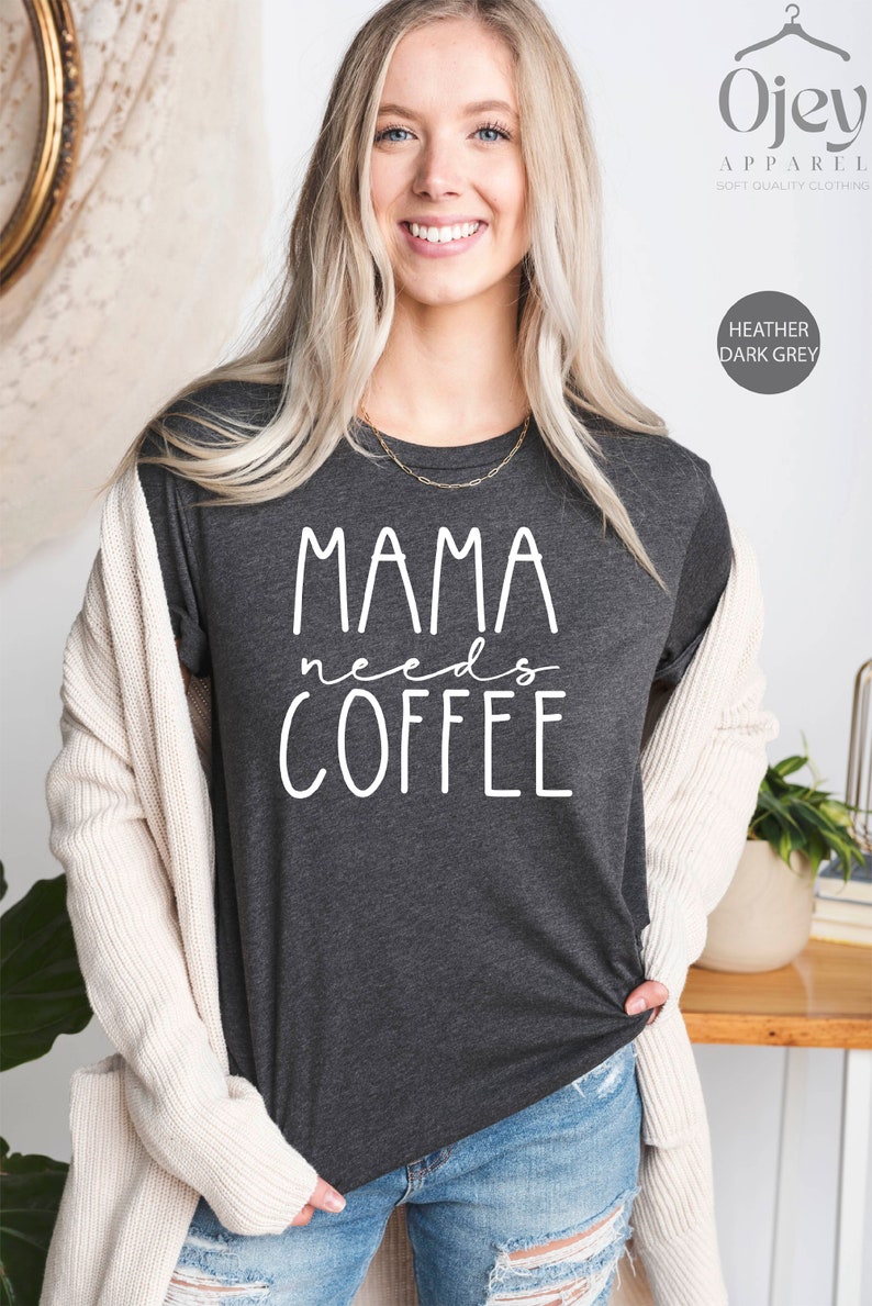 May include: A dark heather grey t-shirt with the text "Mama needs coffee" in white lettering. The shirt is being worn by a person with long blonde hair.