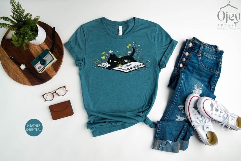 Puede incluir: Una camiseta azul turquesa con un gato negro leyendo un libro rodeado de flores. El texto "HEATHER DEEP TEAL" es visible en la esquina inferior izquierda de la imagen.