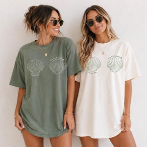 May include: Two oversized crewneck t-shirts, one in sage green and one in cream, each featuring two white line-art seashell graphics on the chest. Below the shells, the text reads "Emma's Bachelorette" in a thin, cursive script font.