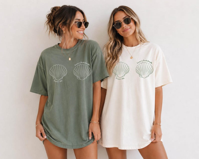 May include: Two oversized crewneck t-shirts, one in sage green and one in cream, each featuring two white line-art seashell graphics on the chest. Below the shells, the text reads "Emma's Bachelorette" in a thin, cursive script font.