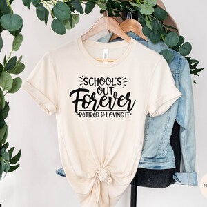 Puede incluir: Una camiseta beige con el texto "School's Out Forever - Retired & Loving It" impreso en negro.