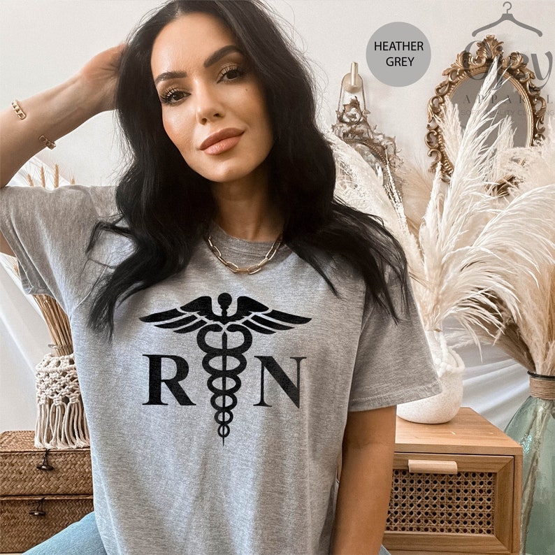 May include: Heather grey t-shirt with a black medical symbol and the letters "RN" printed on the front.