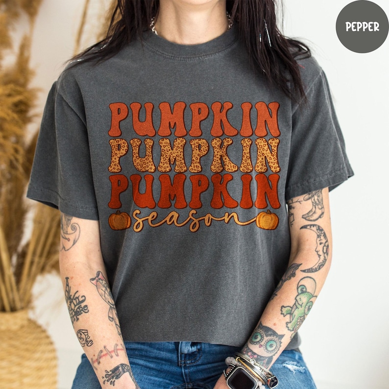 May include: Dark gray t-shirt with the text "PUMPKIN PUMPKIN PUMPKIN season" in orange and leopard print lettering. The shirt has a crew neck and short sleeves. Two small orange pumpkins are on the bottom of the shirt.
