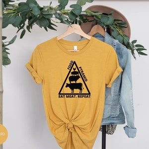 May include: A mustard yellow t-shirt with a black graphic of a food pyramid featuring a cow, pig, and chicken. The text "Food Pyramid" is written on the side of the pyramid and the text "Eat Meat. Repeat." is written below the pyramid.