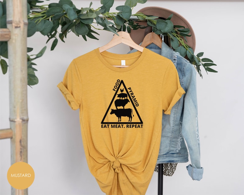 May include: A mustard yellow t-shirt with a black graphic of a food pyramid featuring a cow, pig, and chicken. The text "Food Pyramid" is written on the side of the pyramid and the text "Eat Meat. Repeat." is written below the pyramid.