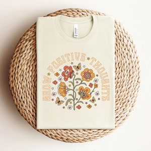 May include: A cream colored t-shirt with a floral design and the text "Grow Positive Thoughts" in a circle around the flowers.