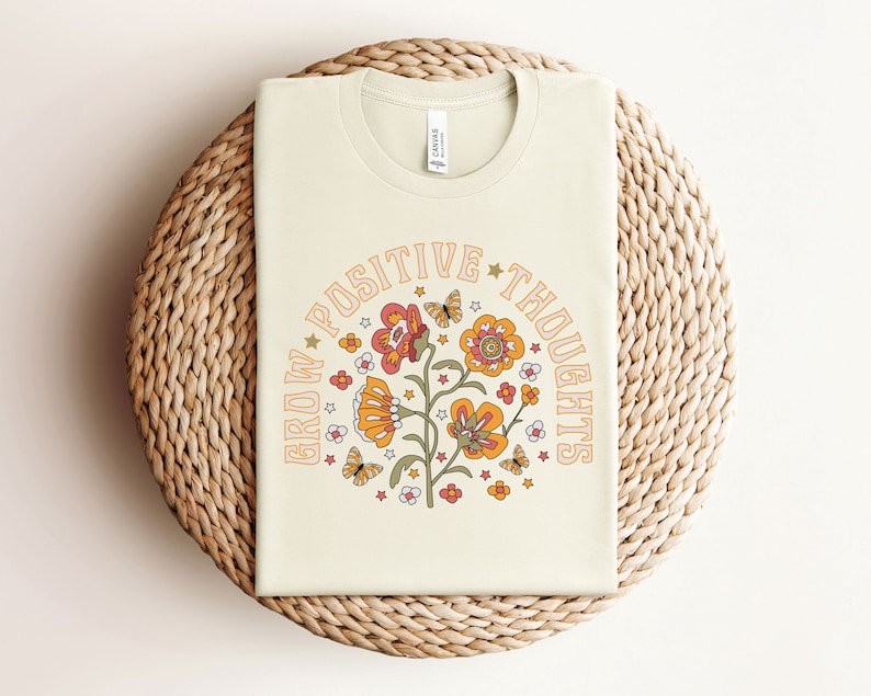 May include: A cream colored t-shirt with a floral design and the text "Grow Positive Thoughts" in a circle around the flowers.