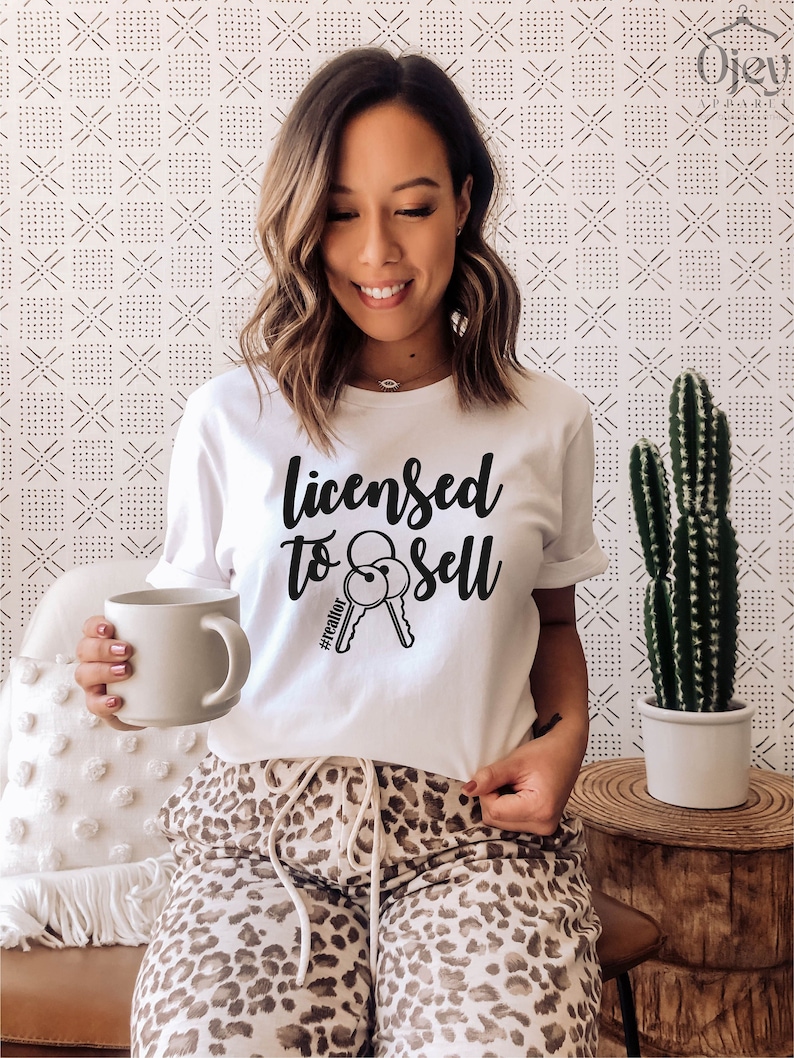 Pode incluir: Camiseta branca com texto preto que diz "Licensed to Sell #realtor" e um gr&aacute;fico de duas chaves. A pessoa que est&aacute; usando a camiseta est&aacute; sentada em um sof&aacute; com estampa de leopardo.
