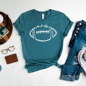 Peut inclure: Un t-shirt en jersey chin&eacute; bleu sarcelle avec un motif blanc de ballon de football et le texte "game day".