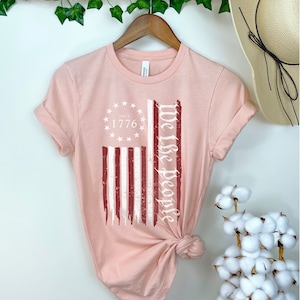 May include: A pink t-shirt with a distressed red, white, and blue American flag design. The text "We the People" is printed vertically on the right side of the flag. The text "Since 1776" is printed inside a circle of stars above the flag.
