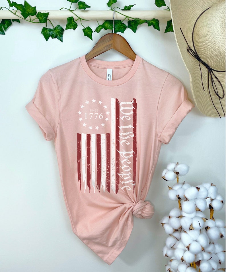 May include: A pink t-shirt with a distressed red, white, and blue American flag design. The text "We the People" is printed vertically on the right side of the flag. The text "Since 1776" is printed inside a circle of stars above the flag.