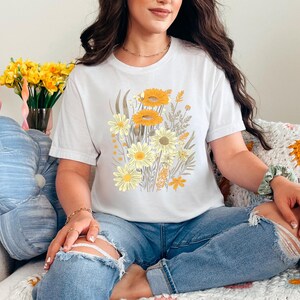 K&ouml;nnte beinhalten: Ein wei&szlig;es T-Shirt mit einem Blumendruck mit gelben und wei&szlig;en Blumen. Das Shirt wird von einer Person getragen, die auf einer geh&auml;kelten Decke sitzt.