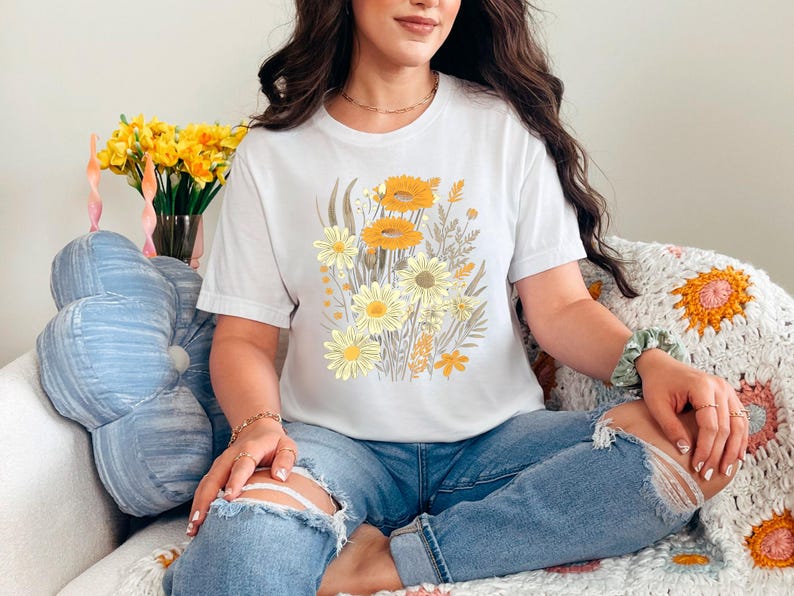 K&ouml;nnte beinhalten: Ein wei&szlig;es T-Shirt mit einem Blumendruck mit gelben und wei&szlig;en Blumen. Das Shirt wird von einer Person getragen, die auf einer geh&auml;kelten Decke sitzt.