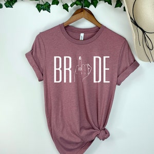 May include: A mauve t-shirt with the word "BRIDE" printed in white. The letter "I" is replaced with a stylized image of a hand with a ring on the middle finger.