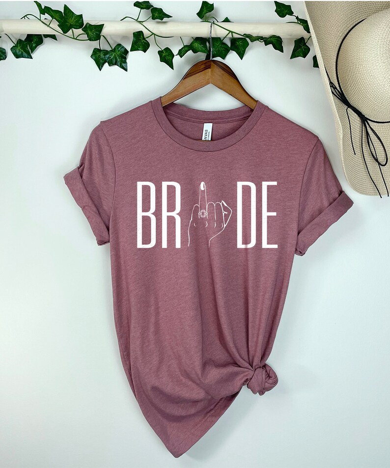 May include: A mauve t-shirt with the word "BRIDE" printed in white. The letter "I" is replaced with a stylized image of a hand with a ring on the middle finger.
