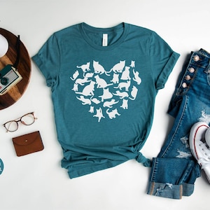 May include: A teal blue t-shirt with a white heart-shaped design made up of cat silhouettes. The shirt is laid flat on a white surface with a pair of glasses, a brown wallet, a wooden tray with a camera and a plant, and a pair of blue jeans and white sneakers.