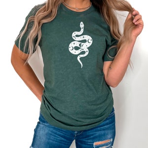 May include: A dark green t-shirt with a white snake design. The snake is decorated with stars, moons, and other celestial elements.