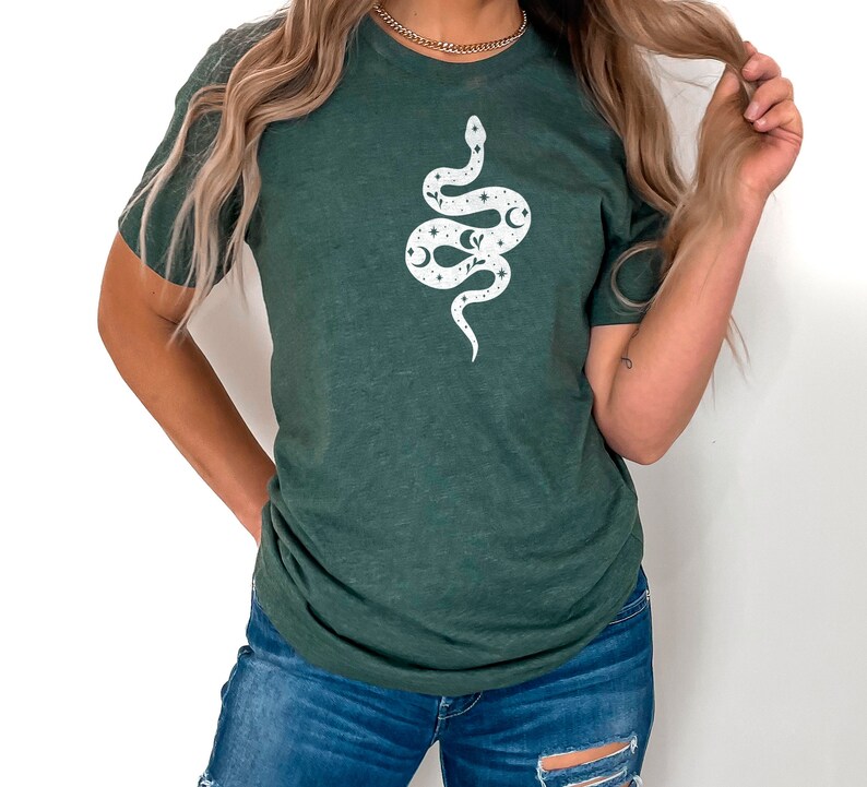 May include: A dark green t-shirt with a white snake design. The snake is decorated with stars, moons, and other celestial elements.