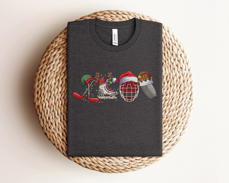 May include: A dark gray t-shirt with a Christmas-themed hockey design. The design features a hockey stick, skates, a hockey helmet, and a Santa hat.