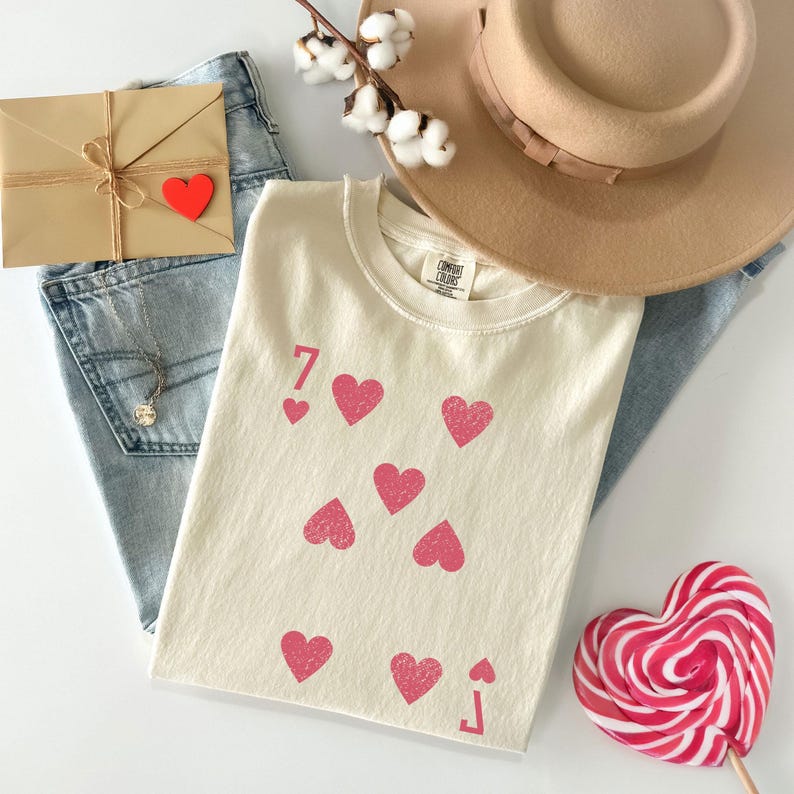 May include: Cream-colored t-shirt with a playing card design featuring the number 7 and pink hearts. The shirt is paired with blue jeans, a tan hat, a heart-shaped lollipop, and a small package tied with twine and a red heart.