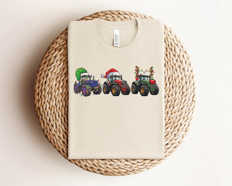 Peut inclure: Un t-shirt beige avec un motif de No&euml;l repr&eacute;sentant trois tracteurs d&eacute;cor&eacute;s de lumi&egrave;res et d'accessoires festifs. Les tracteurs sont bleu, rouge et vert.
