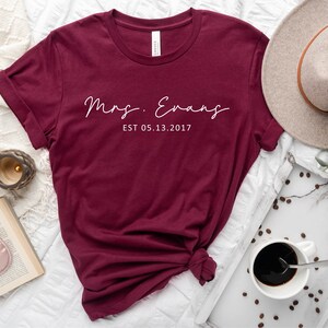 May include: Maroon t-shirt with white text that reads "Mrs. Evans EST 05.13.2017".