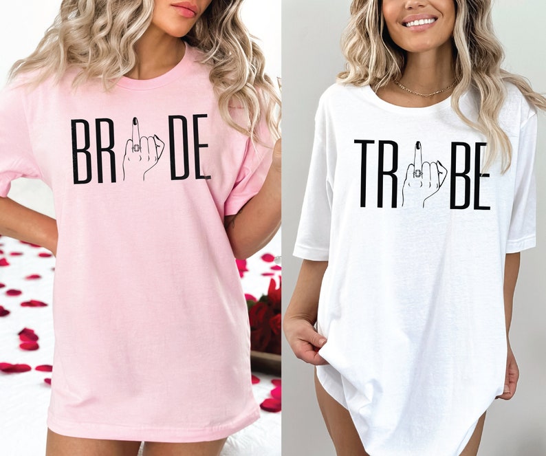 May include: Two t-shirts, one pink and one white, with the words "BRIDE" and "TRIBE" printed on them. Both shirts feature a stylized hand with a ring on the middle finger giving a middle finger gesture.