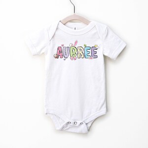 May include: White baby onesie with the name "AUBREE" in colorful, pastel letters. The design includes Easter-themed graphics like a bunny and eggs. The onesie is hanging on a white hanger.