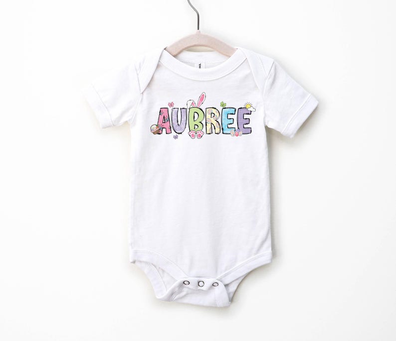 May include: White baby onesie with the name "AUBREE" in colorful, pastel letters. The design includes Easter-themed graphics like a bunny and eggs. The onesie is hanging on a white hanger.