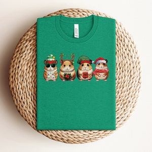May include: Green t-shirt with a graphic of four hamsters wearing Christmas hats and holding gifts. The hamsters are wearing Santa hats, elf hats, and reindeer antlers.
