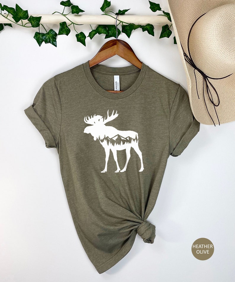 May include: Heather olive green t-shirt featuring a white moose silhouette with a mountain and forest design inside. The shirt is hanging on a wooden hanger. The text "HEATHER OLIVE" is in a circle on the bottom right.