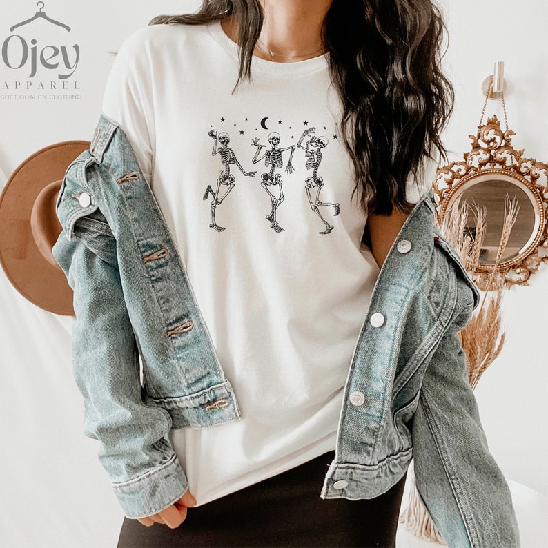 May include: White t-shirt featuring a graphic of three skeletons dancing under a crescent moon and stars.
