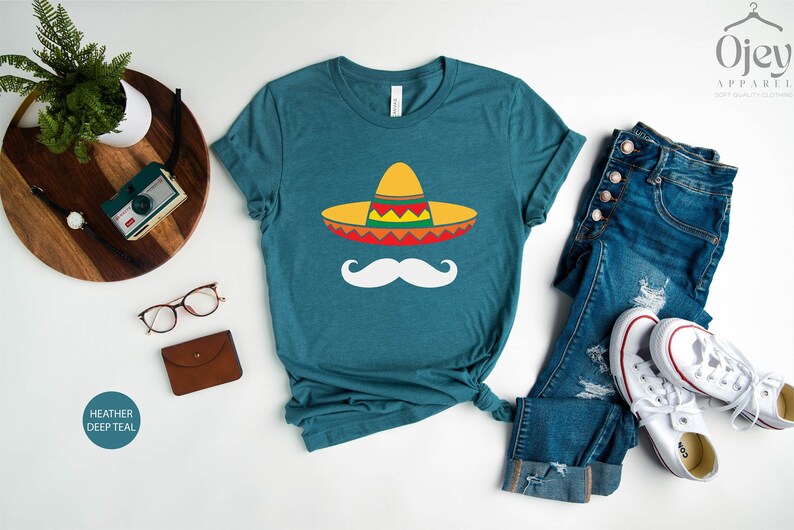 May include: A teal blue t-shirt with a graphic of a sombrero and a mustache. The text "Heather Deep Teal" is visible in the lower left corner of the image.