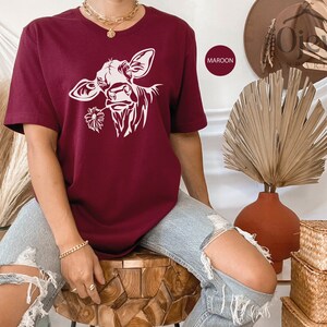 May include: A maroon t-shirt with a white silhouette of a cow's head with a flower in its mouth. The word "MAROON" is written in white on a black rectangle.