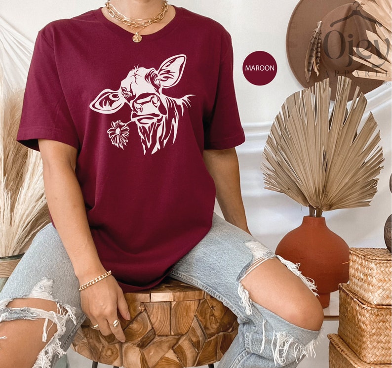 May include: A maroon t-shirt with a white silhouette of a cow's head with a flower in its mouth. The word "MAROON" is written in white on a black rectangle.