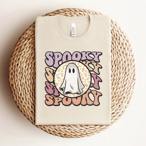 Puede incluir: Una camiseta beige con un gr&aacute;fico de un fantasma en un c&iacute;rculo con la palabra "SPOOKY" en una fuente retro alrededor del c&iacute;rculo.