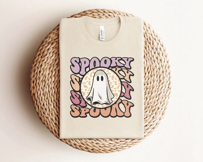 Puede incluir: Una camiseta beige con un gr&aacute;fico de un fantasma en un c&iacute;rculo con la palabra "SPOOKY" en una fuente retro alrededor del c&iacute;rculo.